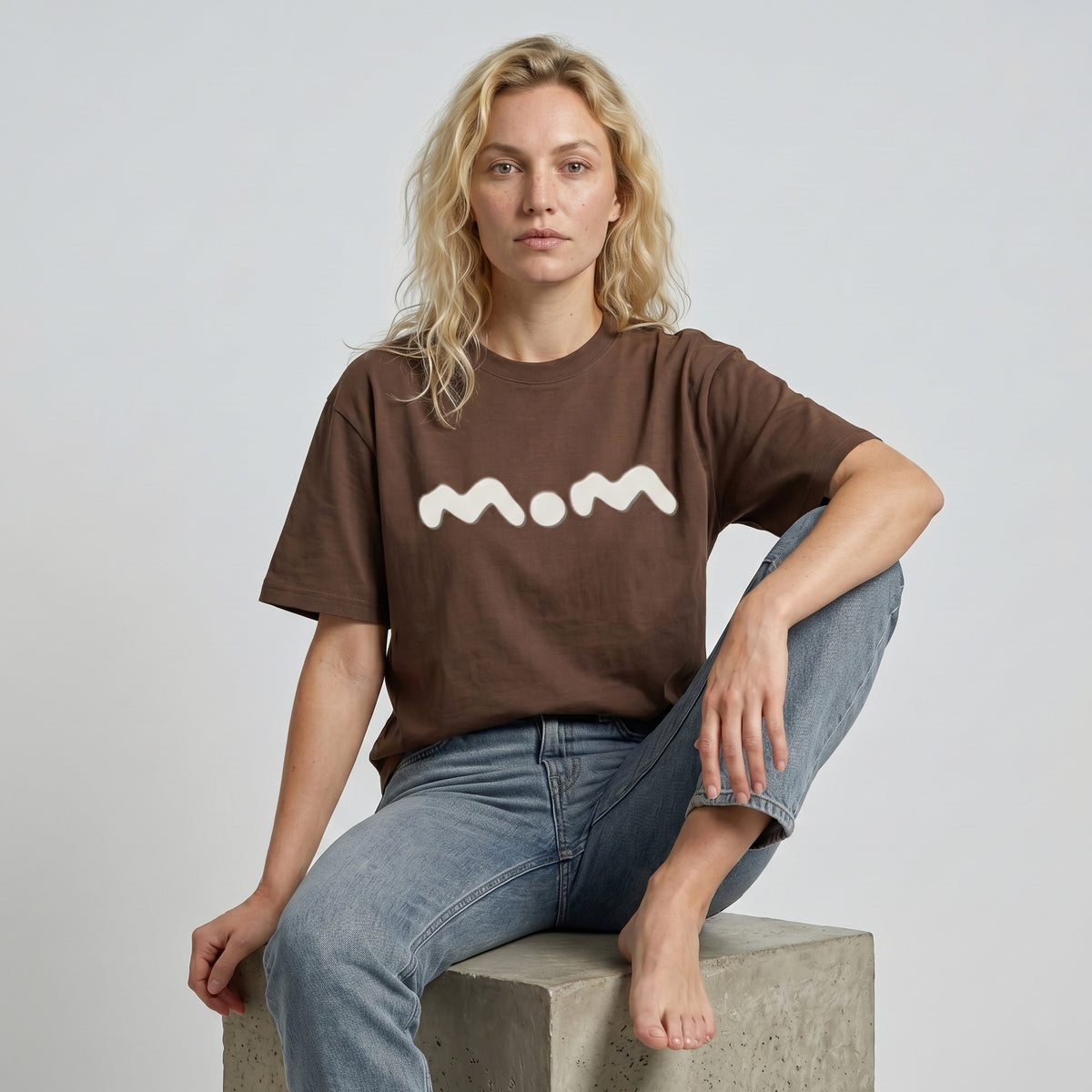 Person wearing a brown t-shirt with 'mom' printed on it, sitting on a concrete block against a plain background.