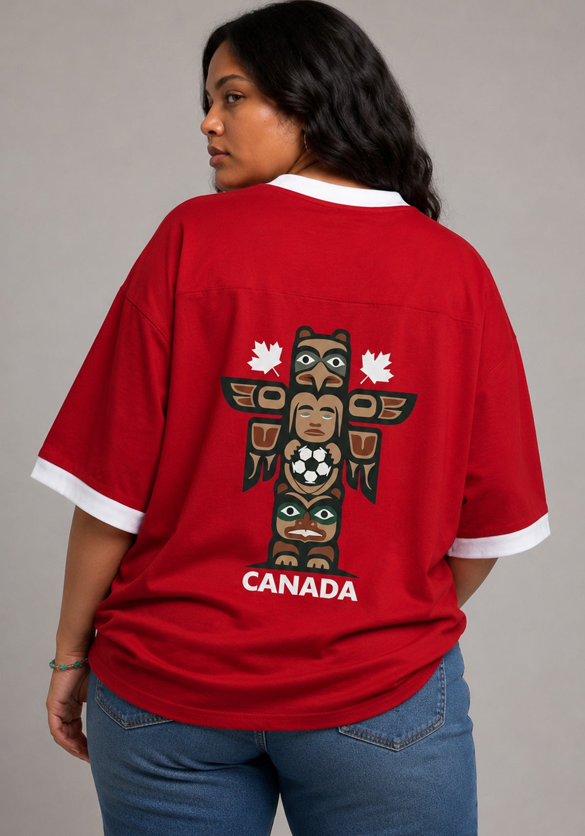 Back view of a model wearing the red Canada jersey, showcasing a graphic of an Indigenous-inspired totem pole holding a soccer ball with the word "CANADA" printed in white below.