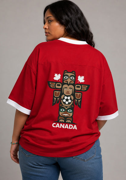 Back view of a model wearing the red Canada jersey, showcasing a graphic of an Indigenous-inspired totem pole holding a soccer ball with the word "CANADA" printed in white below.