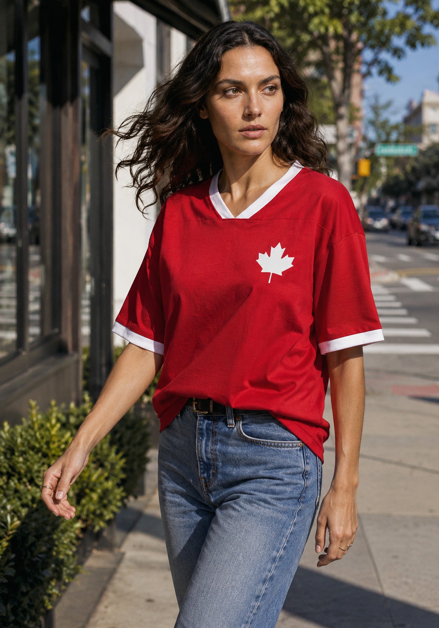 A woman walking on a city street wearing a red and white Blokecore style Canada jersey tucked into blue jeans.
