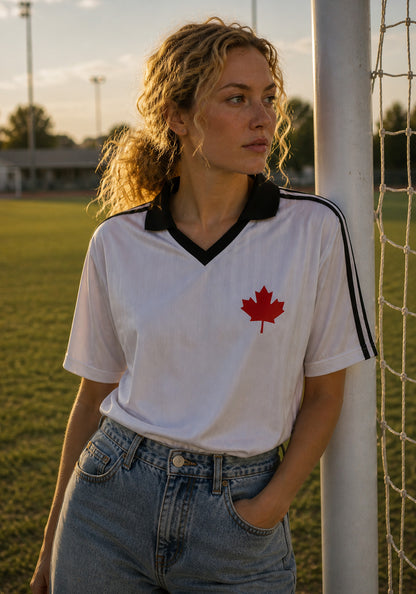 A lifestyle shot of a woman in a white Canadian soccer jersey and light-wash denim, leaning against a white goal post on a soccer field during golden hour.