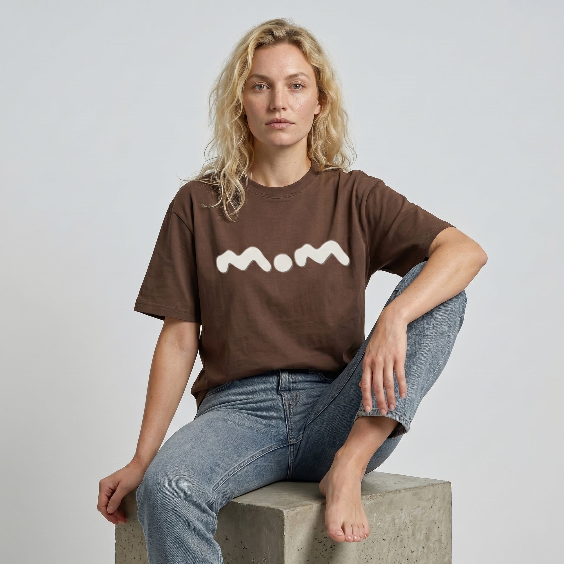 Person wearing a brown t-shirt with 'mom' printed on it, sitting on a concrete block against a plain background.