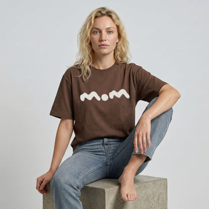 Person wearing a brown t-shirt with 'mom' printed on it, sitting on a concrete block against a plain background.